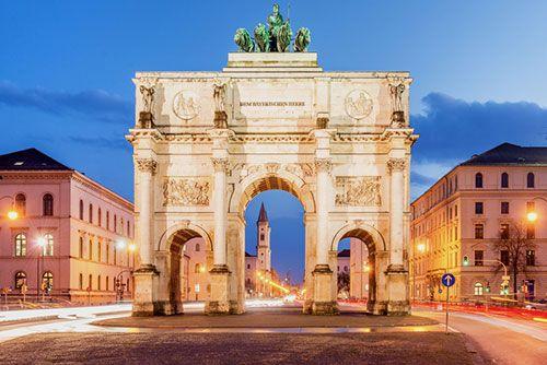 Siegestor in München am Abend, beleuchtet mit vorbeifahrendem Verkehr.
